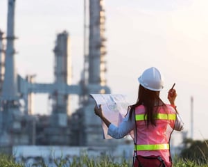 An engineer wearing a hard hat and high-visibility vest stands outdoors, holding blueprints and a walkie-talkie while facing an industrial facility in the distance, representing on-site operations, infrastructure planning, and industrial project oversight.
