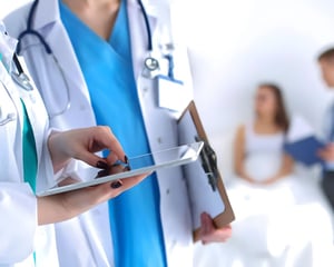 Healthcare professionals in lab coats review information on a tablet and clipboard, while a pregnant patient consults with a clinician in the background, representing coordinated, technology-enabled prenatal or clinical care.