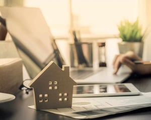 A small wooden house model sits on financial documents beside a tablet and laptop on a desk, representing real estate operations, property management, and technology-supported business growth.