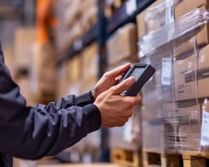 A warehouse worker scans stacked boxes on shelving with a handheld device, representing inventory management, logistics operations, and technology-enabled supply chain processes.