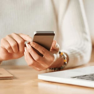 A woman sitting at a desk wiht a computer in front of her and using a mobile phone