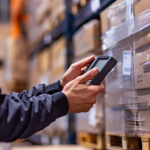 A warehouse worker scans stacked boxes on shelving with a handheld device, representing inventory management, logistics operations, and technology-enabled supply chain processes.