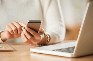 A woman sitting at a desk wiht a computer in front of her and using a mobile phone