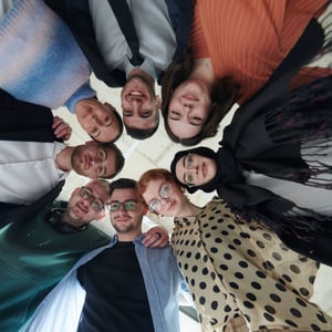 A diverse group of colleagues stands in a circle, looking down at the camera and smiling, creating a sense of teamwork and inclusion in a modern workplace setting.