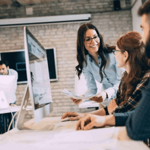 A female technical expert is standing and looking at another female sitting at the desk with a computer screen in front of her.