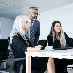 Two women at a desk with a man standing behind and viewing a computer screen